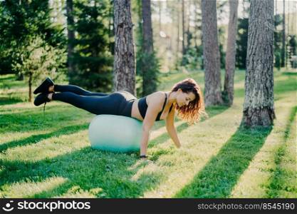Outdoor shot of sporty young woman leans at fitness ball keeps balance and does gymnastic exercises wears cropped top leggings sneakers has workout in forest poses on green grass near trees.