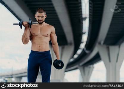 Outdoor shot of strong muscular man raises barbells and trains muscles stands under bridge has aim to have healthy body shape, has fitness training or physical workout. Athletic bodybuilder.