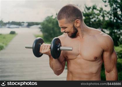 Outdoor shot of strong muscular man with naked torso lifts heavy barbell and trains muscles has workout for keeping healthy fit. Athletic guy demonstrates determination motivates to go in for sport