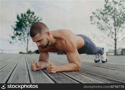 Outdoor shot of strong sportsman stands in plank pose, puts all efforts in keeping fit and healthy. Athlete man with concentrated expression, works out in open air. Bodybuilder makes press exercise