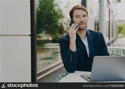 Outdoor shot of successful man executive worker calls someone via smartphone consults customer by phone sits at table in cafe waits for order uses laptop for work online. Manager holds interview. Outdoor shot of successful man executive worker calls someone via smartphone