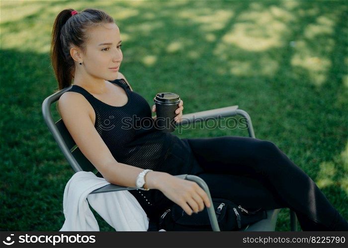 Outdoor shot of thoughtful dark haired beautiful female dressed in sports clothes drinks takeaway coffee spends free time in open air poses on chair at garden enjoys good weather breathes fresh air