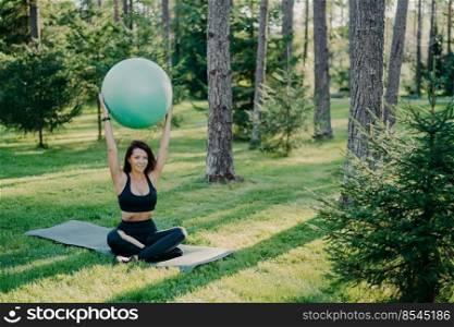 Outdoor shot of young brunette female practices yoga in green park holds fitness ball over head wears cropped top and leggings sits on karemat, breathes fresh air deeply, leads active lifestyle