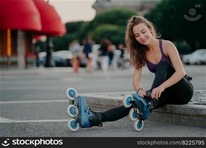 Outdoor shot of young sporty woman in sportswears puts on roller blades poses on road going to ride rollers during sunny day ties shoelaces has good mood. Recreation hobby lifestyle concept.