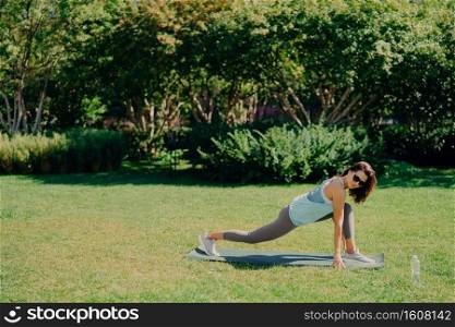 Outdoor sport concept. Healthy fit energetic young woman does fitness exercises on fitness mat dressed in active wear drinks fresh water wears sunglasses poses on green grass. Morning workout.