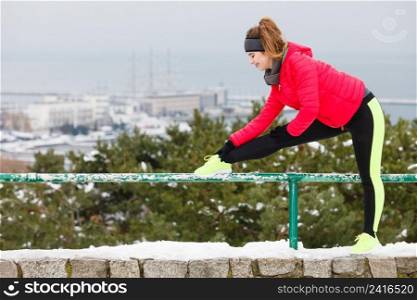 Outdoor sport exercises, sporty outfit ideas. Woman wearing warm sportswear training exercising stretching legs outside during winter. Cityscape panorama in background. Woman wearing sportswear exercising during winter