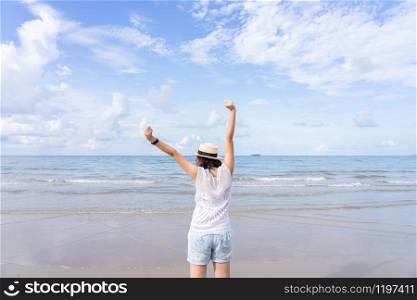 Outdoor summer portrait of Young Asian woman wearing stylish hat and clothes standing on the beach, enjoying looking view of sea with blue sky on summer vacation.