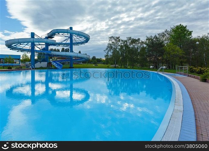 Outdoor swimming pool view with slide and trees reflection on water surface.