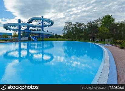 Outdoor swimming pool view with slide and trees reflection on water surface.