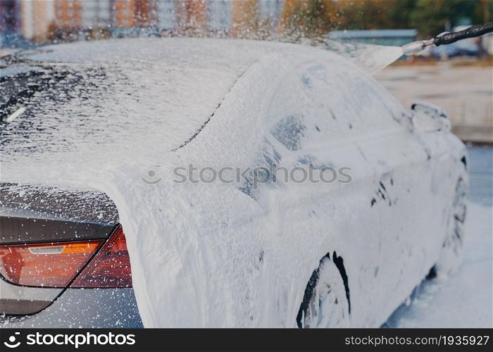 Outdoor vehicle washing. Cleaning process of dirty car with high pressure washer at self-service station, luxury auto in white soap snow foam. Car in white soap foam during cleaning with high pressure washer at carwash station