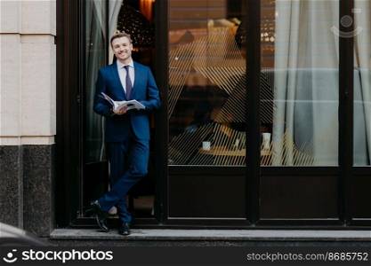 Outdoor view of handsome attractive male executive or CEO in formal suit, stans crossed legs near cozy cafeteria, holds popular journal in hands, has positive expression and stubble. People, lifestyle
