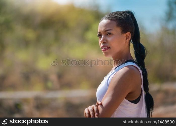 Outdoors portrait of beautiful African Female athlete standing with arms crossed against nature background. Black woman looking away at the park, folded arms and confident expression. Sporty girl doing workout. Healthy lifestyle.