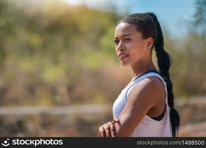 Outdoors portrait of beautiful African Female athlete standing with arms crossed against nature background. Black woman looking away at the park, folded arms and confident expression. Sporty girl doing workout. Healthy lifestyle.