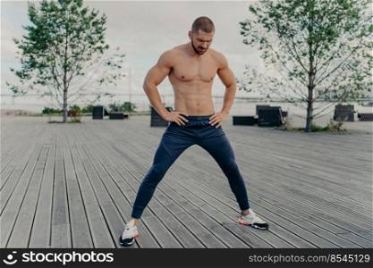 Outside of athlete guy does sport exercises in fresh air, stands with naked torso, has muscular body and stands outside. Active unshaven guy in good physical shape, has regular workout in morning