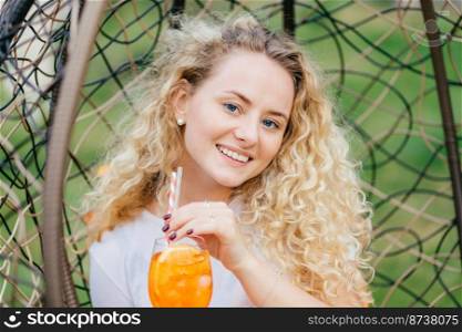 Outside shot of good looking curly female with friendy exprression, drinks summer fresh cocktail, poses in hanging chair, has positive smile, breathes fresh air. People, rest, lifestyle concept