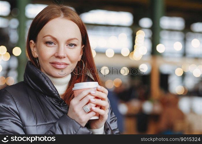Outside shot of satisfied attractive female model with healthy skin, brown hair, holds takeaway coffee, enjoys good drink, dressed in jacket, waits for someone, has coffee break, looks at camera