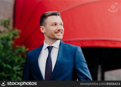 Outside view of handsome young male entrepreneur in formal suit, looks aside with glad expression, notices something pleasant, likes his work, stands outdoor. People, business, lifestyle concept