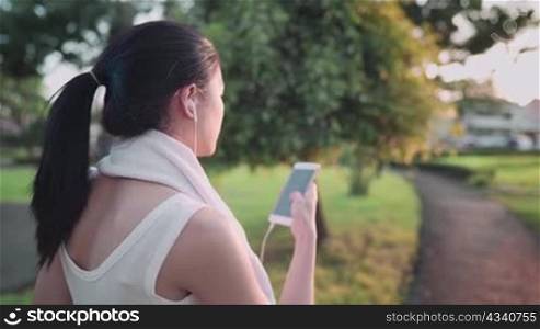 Over shoulder shot asian woman using smartphone while walking down on the walking path at outdoor garden during warm sunset hour, relaxing after workout, wiping sweat from face, technology lifestyle