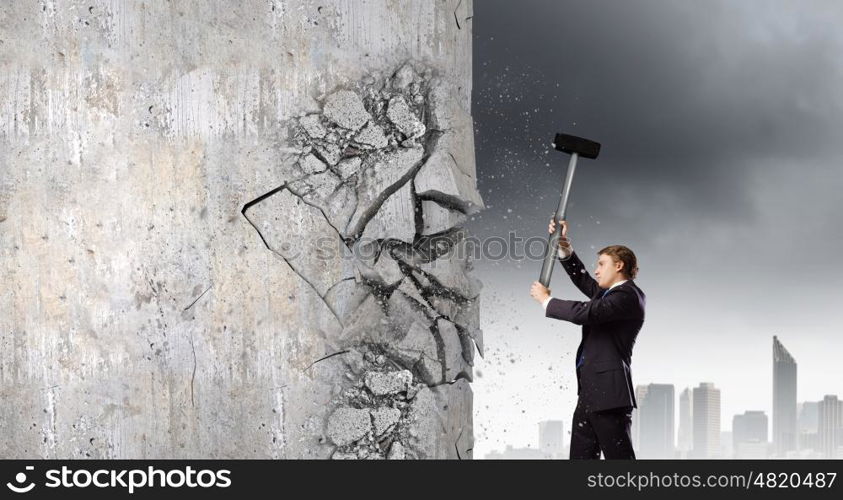 Overcoming challenges. Young determined businessman crashing wall with hammer