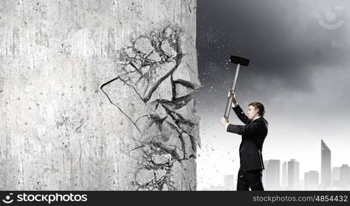 Overcoming challenges. Young determined businessman crashing wall with hammer