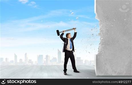 Overcoming challenges. Young determined businessman crashing wall with hammer