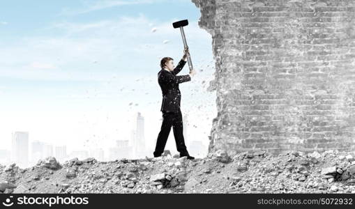 Overcoming challenges. Young determined businessman crashing wall with hammer