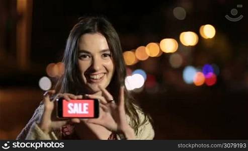 Overexcited shopaholic female holding smartphone with red screen and sale text ad, gesturing and smiling on night city street. Overjoyed beautiful woman showing mobile phone screen with sale sign to camera at night over streelights bokeh background.