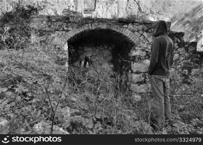 Overgrown plants and hooded man next to arched recess of rural ruin.