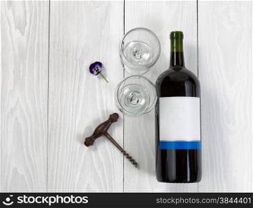 Overhead angled view of a large bottle of red wine, drinking glasses, flower and antique corkscrew on white wooden boards.