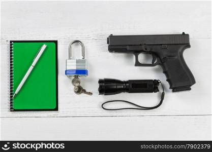 Overhead angled view of security items consisting of pistol, flashlight, pen, notepad and padlock with keys on desktop.