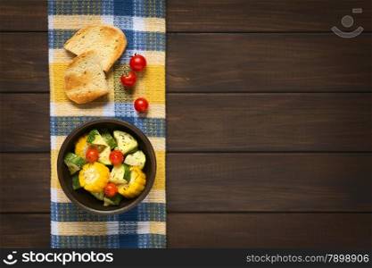 Overhead shot of a bowl of baked vegetables of sweet corn, zucchini, cherry tomato with thyme, toasted bread slices above, photographed on dish towel on dark wood with natural light