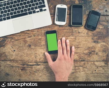 Overhead shot of a male hand using a smart phone with a green screen on a wooden table. There is a laptop and other smart phones on the table too.