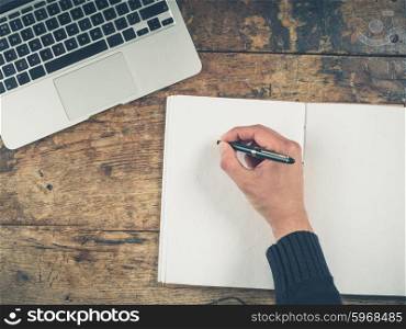 Overhead shot of a male hand writing in a notepad. There is a laptop on the table as well.