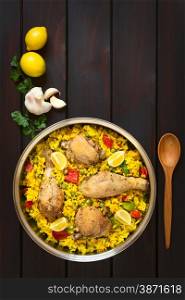 Overhead shot of a pot of chicken paella, a traditional Valencian (Spanish) rice dish made of rice, chicken, peas, capsicum and served with lemon, photographed on dark wood with natural light
