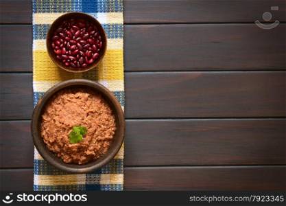 Overhead shot of a rustic bowl of homemade red kidney bean spread garnished with fresh coriander leaf, a bowl of raw kidney beans above, photographed on dark wood with natural light