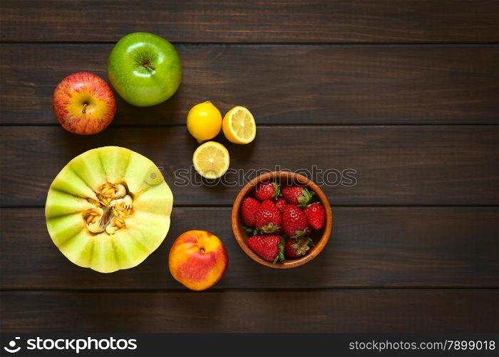 Overhead shot of a variety of fresh fruits (honeydew melon, nectarine, apple, strawberry, plum, lemon), photographed on dark wood with natural light