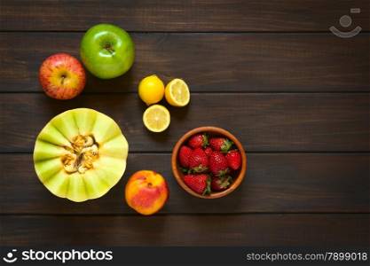 Overhead shot of a variety of fresh fruits (honeydew melon, nectarine, apple, strawberry, plum, lemon), photographed on dark wood with natural light