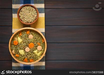 Overhead shot of a wooden bowl of lentil soup made with potato, carrot, onion and sausage slices, with small bowl of raw lentils above, photographed on kitchen towel on dark wood with natural light