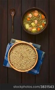 Overhead shot of a wooden bowl of raw lentils and a rustic bowl of lentil soup made with potato, carrot, onion and sausage slices, photographed on dark wood with natural light