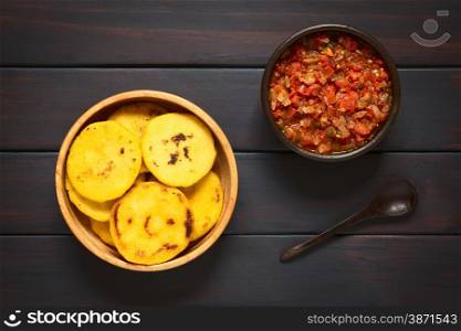 Overhead shot of arepas in wooden bowl with Colombian hogao sauce (tomato and onion cooked) and a spoon. Arepas are made of yellow or white corn meal and are traditionally eaten in Colombia and Venezuela. Photographed on dark wood with natural light.. Colombian Arepa with Hogao Sauce