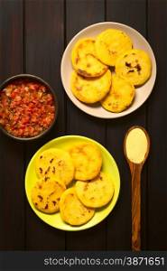 Overhead shot of arepas on plates with Colombian hogao sauce (tomato and onion cooked) in bowl. Arepas are made of yellow or white corn meal and are traditionally eaten in Colombia and Venezuela. Photographed on dark wood with natural light.. Colombian Arepa with Hogao Sauce