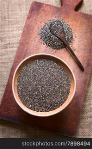 Overhead shot of chia seeds (lat. Salvia hispanica) in clay bowl photographed on wooden board with natural light. Chia seeds are considered a superfood containing proteins, omega fats, minerals and antioxidants (Selective Focus, Focus on the seeds in the bowl)