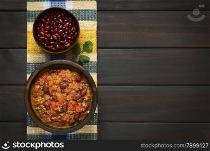 Overhead shot of chili con carne and dried kidney beans in bowls on kitchen towel, photographed on dark wood with natural light
