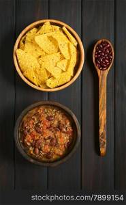 Overhead shot of chili con carne and tortilla chips in bowls with dried kidney beans on wooden spoon, photographed on dark wood with natural light