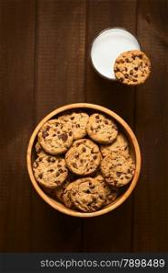 Overhead shot of chocolate chip cookies in wooden bowl and a cookie on the rim of a glass of cold milk, photographed on wood with natural light