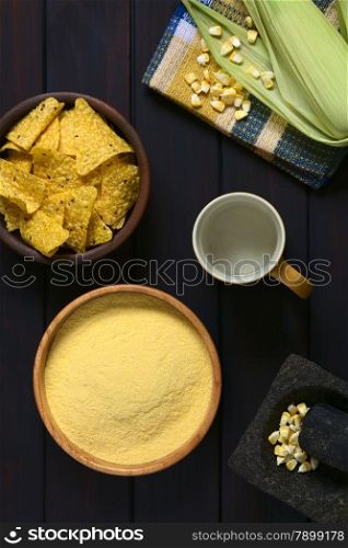Overhead shot of cornmeal in wooden bowl with cob of corn, corn kernels in mortar, cup of water and tortilla chips in bowl, photographed on dark wood with natural light