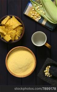 Overhead shot of cornmeal in wooden bowl with cob of corn, corn kernels in mortar, cup of water and tortilla chips in bowl, photographed on dark wood with natural light