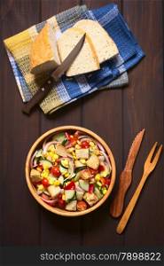 Overhead shot of fresh vegetable salad made of sweet corn, cherry tomato, cucumber, red onion, red pepper, chives with croutons in wooden bowl, photographed on dark wood with natural light