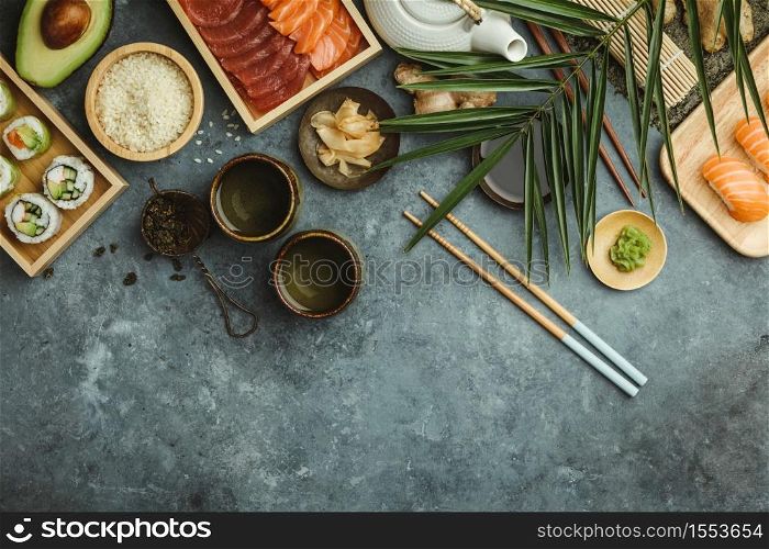 Overhead shot of ingredients for sushi, green tea and prepared maki and nigiri on dark blue background. Raw salmon and tuna pieses, rice, avocado, pickled ginger (gari), raw ginger, wasabi, soy sauce, hopsticks. Asian food background. Space for text. Top view. Overhead shot of ingredients for sushi on dark blue background