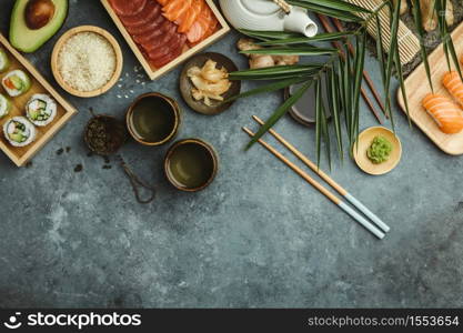 Overhead shot of ingredients for sushi, green tea and prepared maki and nigiri on dark blue background. Raw salmon and tuna pieses, rice, avocado, pickled ginger (gari), raw ginger, wasabi, soy sauce, hopsticks. Asian food background. Space for text. Top view. Overhead shot of ingredients for sushi on dark blue background